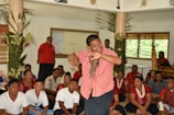 A group of people, some seated on the floor, others standing, are in a room adorned with greenery. A man in the center wearing a red checkered shirt is performing a dance. The audience around him appears engaged and diverse, wearing a variety of colorful outfits. The room has windows with blinds, and the atmosphere seems lively and cultural.