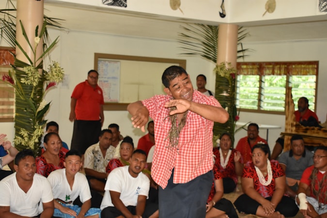A group of people, some seated on the floor, others standing, are in a room adorned with greenery. A man in the center wearing a red checkered shirt is performing a dance. The audience around him appears engaged and diverse, wearing a variety of colorful outfits. The room has windows with blinds, and the atmosphere seems lively and cultural.