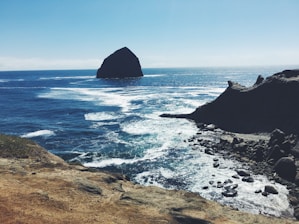 A large rock formation rises out of the ocean, surrounded by blue water and white waves. In the foreground, a rocky coastline stretches into the distance, with rugged cliffs and scattered stones along the shore.