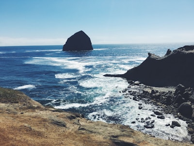 A large rock formation rises out of the ocean, surrounded by blue water and white waves. In the foreground, a rocky coastline stretches into the distance, with rugged cliffs and scattered stones along the shore.