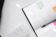 A neatly arranged planner or calendar lies open with various dates filled in with notes and reminders. Several sticky notes in different colors, such as orange and green, are attached to the planner. A black pen is placed diagonally across the open calendar. Nearby, there is a white notepad with handwritten notes detailing a schedule or to-do list.