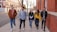 Five young adults walking along a sidewalk next to a red-brick building. They appear to be engaged in light conversation and laughter. There are several trees along the sidewalk and a multi-story building across the street with large windows. It's a bright and clear day.