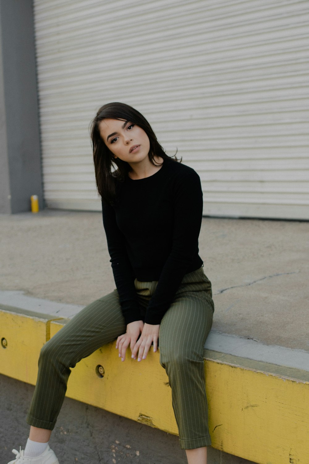 woman in black long-sleeved shirt and grey pinstripe pants sitting near grey shutter door during daytime