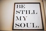 A wooden-framed sign featuring the phrase 'BE STILL MY SOUL' in large, bold black letters on a white background. The sign is leaning against a light-colored wall, creating a calming and minimalist aesthetic.