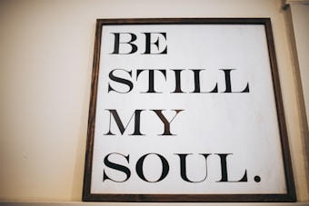 A wooden-framed sign featuring the phrase 'BE STILL MY SOUL' in large, bold black letters on a white background. The sign is leaning against a light-colored wall, creating a calming and minimalist aesthetic.