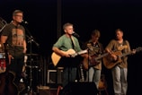 A band of four musicians is performing on stage, with two guitarists and a bass player holding their instruments. A man stands behind a microphone singing, while another musician is off to the side with a microphone stand. The musicians appear focused on their performance, and the lighting centers on them, creating a spotlight effect.