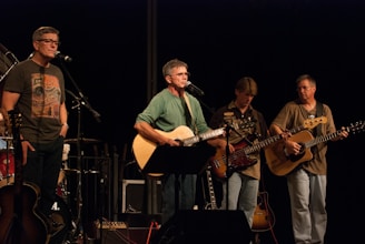 A band of four musicians is performing on stage, with two guitarists and a bass player holding their instruments. A man stands behind a microphone singing, while another musician is off to the side with a microphone stand. The musicians appear focused on their performance, and the lighting centers on them, creating a spotlight effect.