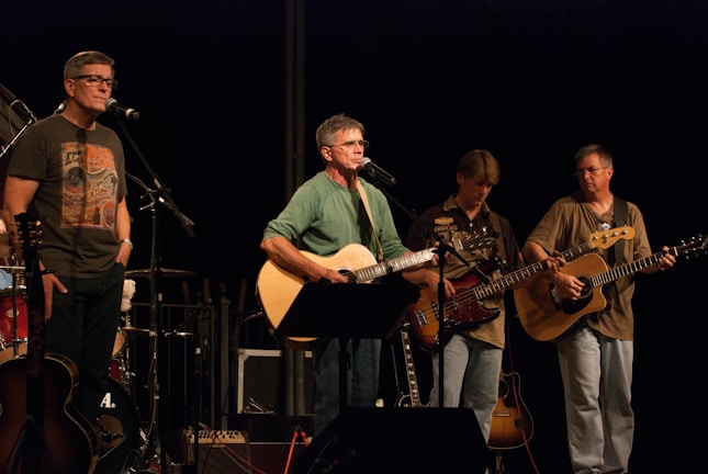 A band of four musicians is performing on stage, with two guitarists and a bass player holding their instruments. A man stands behind a microphone singing, while another musician is off to the side with a microphone stand. The musicians appear focused on their performance, and the lighting centers on them, creating a spotlight effect.