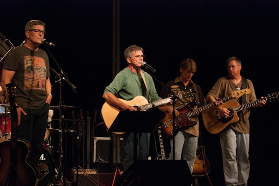 A band of four musicians is performing on stage, with two guitarists and a bass player holding their instruments. A man stands behind a microphone singing, while another musician is off to the side with a microphone stand. The musicians appear focused on their performance, and the lighting centers on them, creating a spotlight effect.