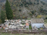 A hillside cemetery with various stone tombs and a small chapel. The tombs are arranged in neat rows, surrounded by lush greenery. Two tall cypress trees stand prominently near the graves. The backdrop consists of a forested hill with autumn colors.