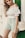 A woman wearing a white off-shoulder top paired with green and white patterned shorts, standing against a green and white background. She carries a straw handbag slung over her shoulder and has long blonde hair.