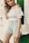 A woman wearing a white off-shoulder top paired with green and white patterned shorts, standing against a green and white background. She carries a straw handbag slung over her shoulder and has long blonde hair.
