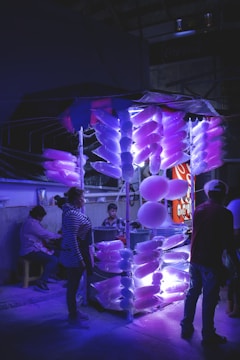 A booth at a night market is illuminated with purple and blue lights, displaying numerous bags of cotton candy. Several people are gathered around the booth, including a vendor and customers. The atmosphere is vibrant yet somewhat moody due to the lighting.