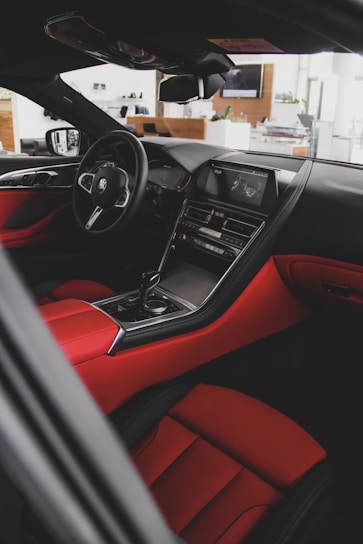 A sleek and modern car interior featuring luxurious red and black leather upholstery. The dashboard is equipped with a high-tech display and control panel. The steering wheel has an emblem, and the overall design conveys elegance and sophistication.