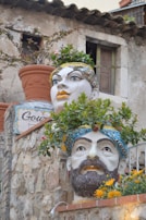 Large ceramic heads with intricate facial features serve as planters, filled with vibrant greenery and flowers. They are displayed against a rustic stone wall with an aged building in the background.