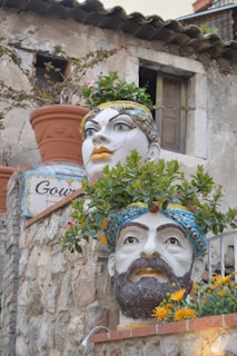 Large ceramic heads with intricate facial features serve as planters, filled with vibrant greenery and flowers. They are displayed against a rustic stone wall with an aged building in the background.