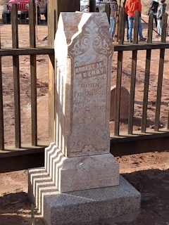 An old, weathered tombstone with intricate carvings and inscriptions is situated in a fenced area. The tombstone reads, 'Robert M. Berry, Born Feb. 3, 1841, Killed by Indians, Apr. 2, 1866.' The background shows a dirt road and several people standing near a wooden fence. A red vehicle is partially visible, adding contrast to the earthy tones of the scene.