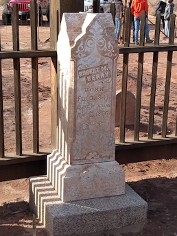 An old, weathered tombstone with intricate carvings and inscriptions is situated in a fenced area. The tombstone reads, 'Robert M. Berry, Born Feb. 3, 1841, Killed by Indians, Apr. 2, 1866.' The background shows a dirt road and several people standing near a wooden fence. A red vehicle is partially visible, adding contrast to the earthy tones of the scene.