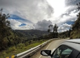 A scenic winding road runs through a mountainous landscape. Tall trees and vegetation line the road, leading towards hills and a partially cloudy sky, creating a sense of movement and adventure. A vehicle is seen on the right side, indicating travel or a road trip.