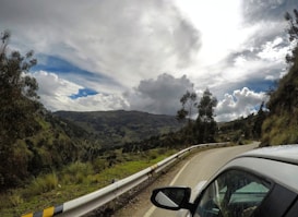A scenic winding road runs through a mountainous landscape. Tall trees and vegetation line the road, leading towards hills and a partially cloudy sky, creating a sense of movement and adventure. A vehicle is seen on the right side, indicating travel or a road trip.