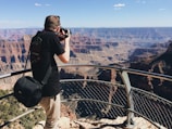 A person is taking a photograph from a viewpoint overlooking a vast canyon landscape. The scene includes rugged cliffs, deep valleys, and a clear blue sky above. The person is leaning on a metal railing, with a camera held to their face, wearing a black t-shirt and beige pants, and carrying a black camera bag.