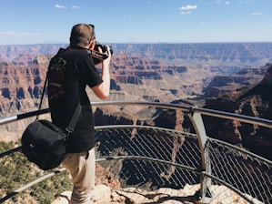 A person is taking a photograph from a viewpoint overlooking a vast canyon landscape. The scene includes rugged cliffs, deep valleys, and a clear blue sky above. The person is leaning on a metal railing, with a camera held to their face, wearing a black t-shirt and beige pants, and carrying a black camera bag.