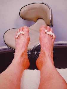 Two feet are propped up against a white towel, with foam separators between the toes. The background features a modern stool with a beige, curved seat on a metal base, set against a light-colored wall.