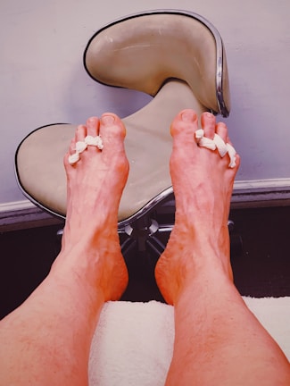 Two feet are propped up against a white towel, with foam separators between the toes. The background features a modern stool with a beige, curved seat on a metal base, set against a light-colored wall.