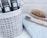 A collection of grooming items including black bottles with the word 'saison' and 'winter' printed on them, placed in a grey and white patterned fabric container. Nearby, a wooden hairbrush with wooden bristles and a bamboo toothbrush with black and white bristles rest on a folded light blue towel.