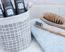 A collection of grooming items including black bottles with the word 'saison' and 'winter' printed on them, placed in a grey and white patterned fabric container. Nearby, a wooden hairbrush with wooden bristles and a bamboo toothbrush with black and white bristles rest on a folded light blue towel.