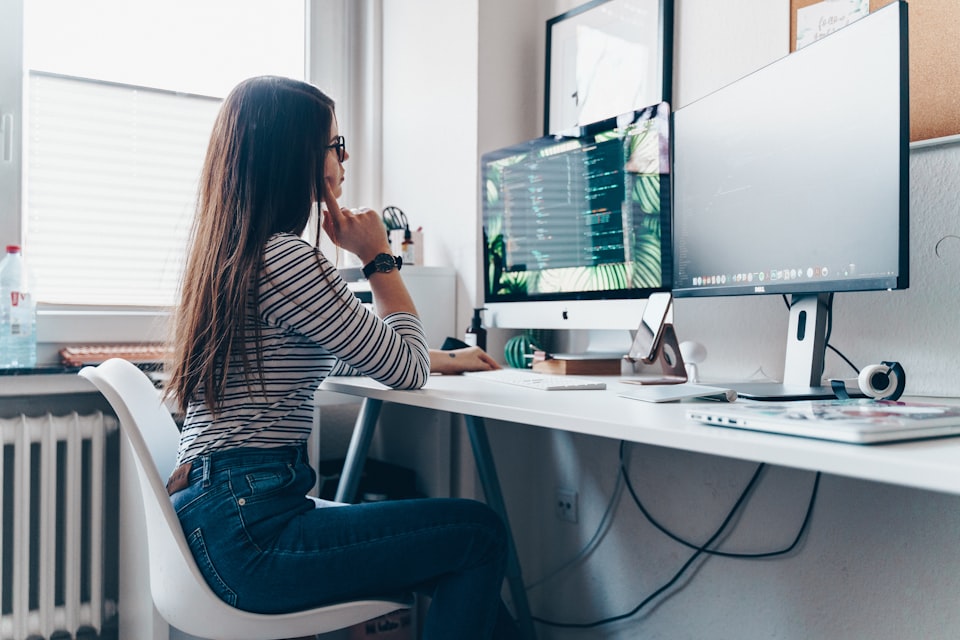 Woman sitting at a white desk while looking at two monitors.
