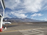 A commercial airplane is parked at an airport tarmac with a mountainous landscape in the background. The scene includes a jet with the LATAM logo, airport staff, and various airport structures. The sky is mostly clear with scattered clouds, providing a serene backdrop.