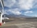 A commercial airplane is parked at an airport tarmac with a mountainous landscape in the background. The scene includes a jet with the LATAM logo, airport staff, and various airport structures. The sky is mostly clear with scattered clouds, providing a serene backdrop.