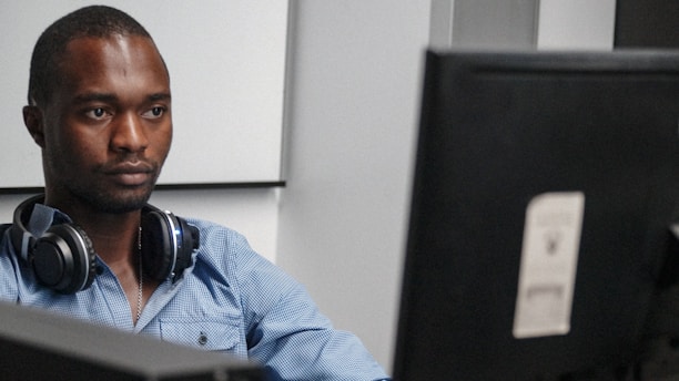 A person wearing a blue shirt and headphones is sitting at a desk focused on a computer monitor. The background features a minimalistic office environment.