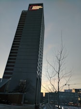 A tall skyscraper with a sign that reads 'Indiana Michigan Power' at the top, set against a clear evening sky. A leafless tree and cranes in the background suggest an urban setting with ongoing construction.