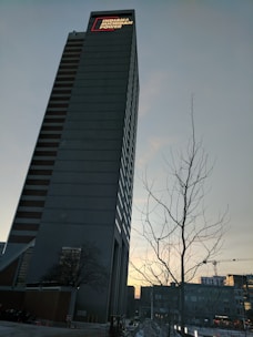 A tall skyscraper with a sign that reads 'Indiana Michigan Power' at the top, set against a clear evening sky. A leafless tree and cranes in the background suggest an urban setting with ongoing construction.