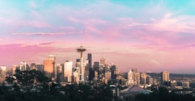 The skyline of a city featuring the iconic Space Needle and a variety of modern skyscrapers is set against a picturesque sky. The sky displays a stunning gradient of pink, purple, and blue hues, suggesting the time is either sunrise or sunset. In the distance, a mountain with a snow-capped peak is visible, adding to the scenic backdrop.