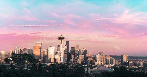 The skyline of a city featuring the iconic Space Needle and a variety of modern skyscrapers is set against a picturesque sky. The sky displays a stunning gradient of pink, purple, and blue hues, suggesting the time is either sunrise or sunset. In the distance, a mountain with a snow-capped peak is visible, adding to the scenic backdrop.