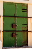 A pair of large green wooden doors are secured with horizontal metal bars and rusted brackets. The doors have vertical panels and are set within a light-colored wall. The metal fixtures and edges of the doors appear aged and weathered.