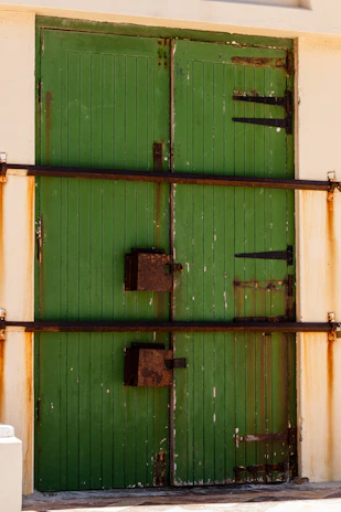 A pair of large green wooden doors are secured with horizontal metal bars and rusted brackets. The doors have vertical panels and are set within a light-colored wall. The metal fixtures and edges of the doors appear aged and weathered.