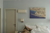 A tidy room with a minimalistic bedside area. There is a neatly made bed with white pillows and a patterned blanket. Above the bed, a striking aerial photograph of a coastal city is mounted on the wall. A closed door is located to the left, and a telephone sits on a small piece of furniture next to the bed. The wall is painted in a soft, muted tone.