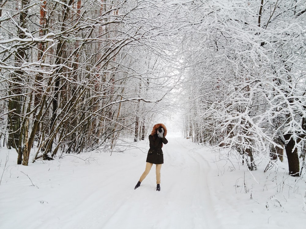 woman in black parka jacket and beige pants standing in the middle of snow covered road surrounded with bare trees