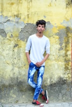 A young person wearing a white long-sleeve shirt and tie-dye blue jeans is standing against an old, peeling yellow and gray wall. They have one hand in their pocket and are casually leaning against the wall. They are also wearing black and red sneakers.