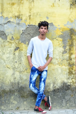 A young person wearing a white long-sleeve shirt and tie-dye blue jeans is standing against an old, peeling yellow and gray wall. They have one hand in their pocket and are casually leaning against the wall. They are also wearing black and red sneakers.