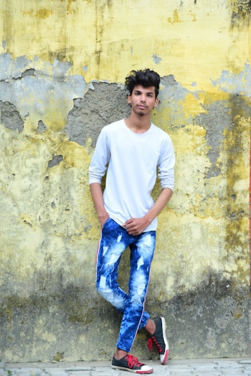A young person wearing a white long-sleeve shirt and tie-dye blue jeans is standing against an old, peeling yellow and gray wall. They have one hand in their pocket and are casually leaning against the wall. They are also wearing black and red sneakers.