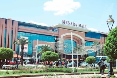 A large, modern shopping mall featuring a grand entrance with palm trees and decorative lighting. The building facade combines glass and a brownish facade. People are walking in and out, with cars parked in front, and outdoor seating visible, suggesting a lively atmosphere.