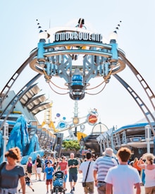 A bustling, futuristic theme park entrance with a sign reading 'Tomorrowland' mounted on a large, elaborate structure. The entrance features various modern, space-themed designs with metallic and colorful elements. Numerous visitors are walking through the area, including families with children, people pushing strollers, and individuals in casual summer attire. The sky is clear and blue, adding to the bright and cheerful atmosphere.