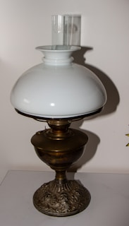 A vintage oil lamp with an ornate metal base and a large white glass shade is set against a plain background. The glass chimney extends upward from the center of the shade.