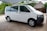 A white van is parked under a canopy. The surroundings include greenery and a thatched-roof building in the background. The area appears to be paved, and the vehicle is facing to the right.