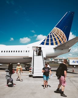 A large commercial airplane is parked on a tarmac under a clear blue sky. People are descending a mobile staircase from the aircraft, carrying bags and wearing casual attire. The airplane has a distinctive blue and gold tail fin design. The background includes airport buildings and a service vehicle.
